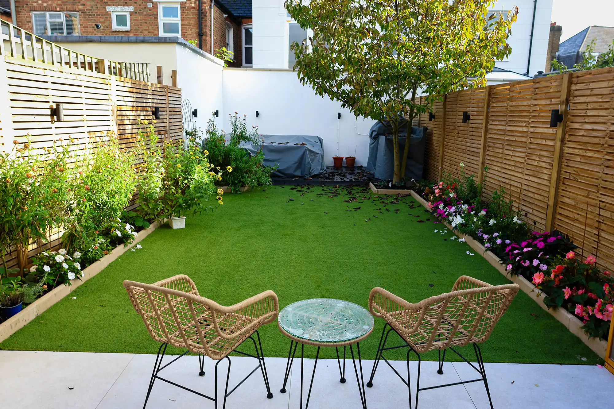 A well-maintained garden with artificial grass, colorful flower beds, and two wicker chairs with a glass table in the foreground.