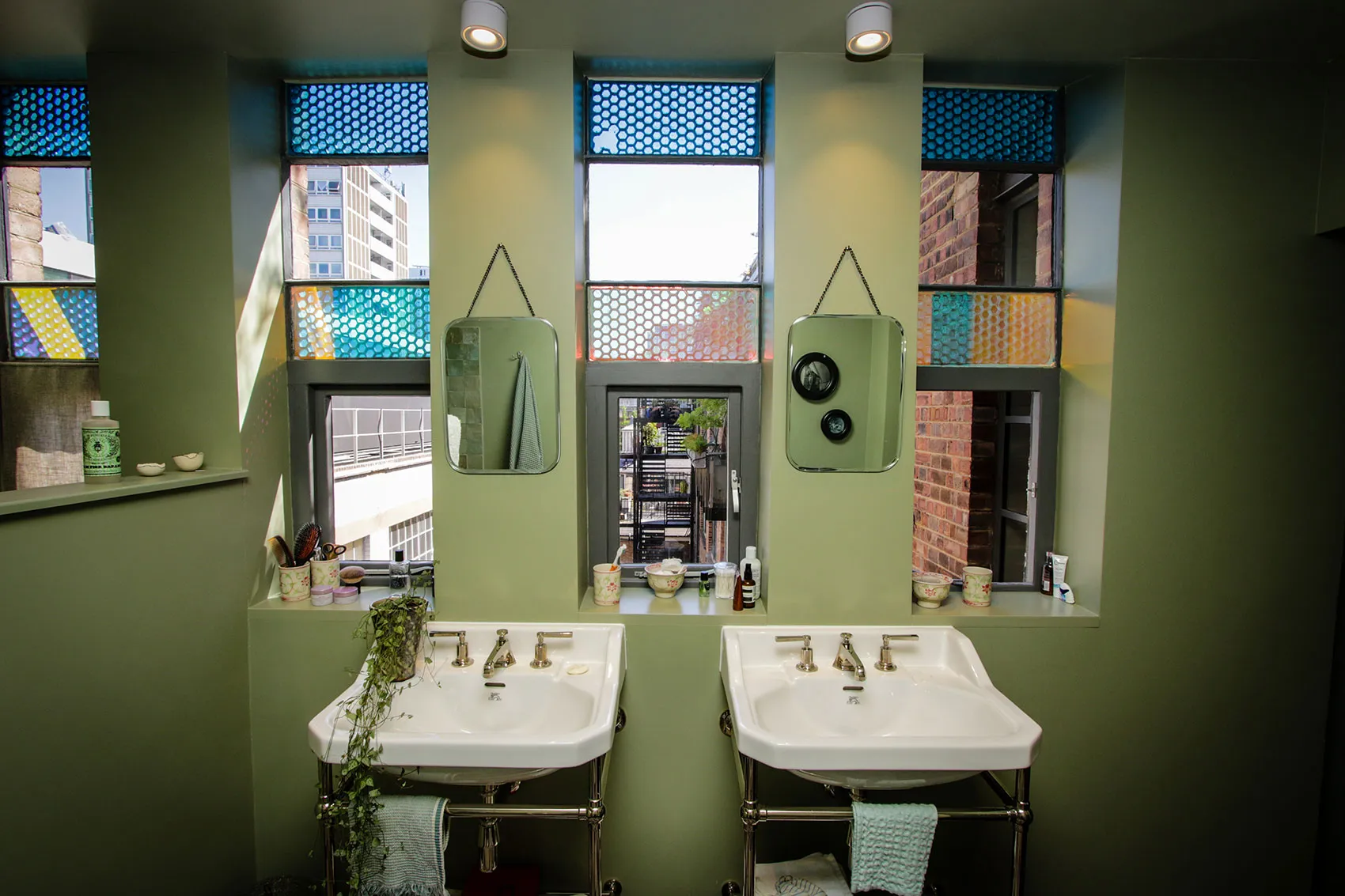 Modern bathroom featuring two white sinks, colorful patterned windows, and green walls. Natural light illuminates the space.