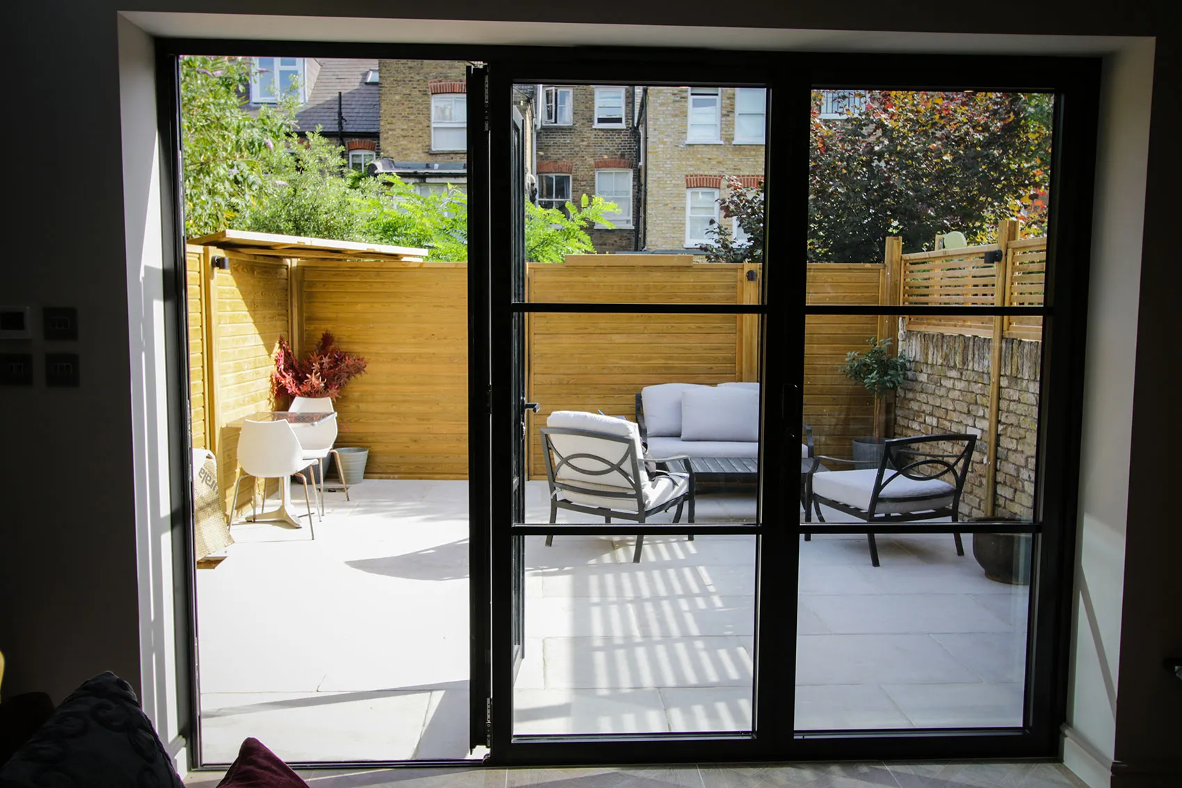 Bright patio view through black-framed doors, featuring seating and plants against a wooden fence backdrop.