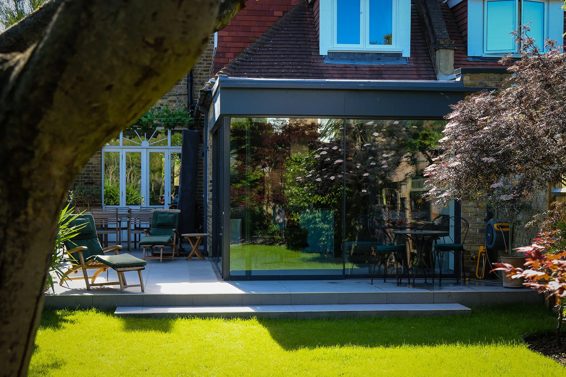 A modern glass extension attached to a brick house, with a green lawn and patio furniture in a lush garden setting.